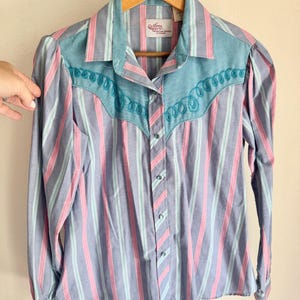 May include: A vintage button-down shirt with vertical stripes in blue, pink, and mint green. The shirt features a turquoise yoke with embroidered detailing and long sleeves. The shirt is hanging on a wooden hanger.