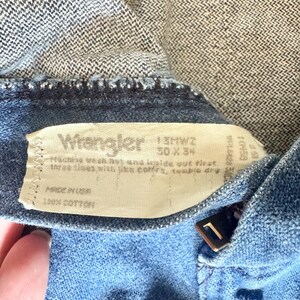 May include: Close-up of a pair of blue denim Wrangler jeans. The jeans have a zipper and a button closure. The label reads "Wrangler" and includes size information: 30 x 34. The jeans are made of 100% cotton.