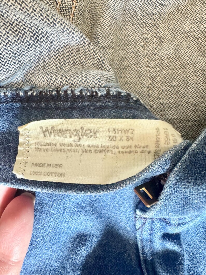 May include: Close-up of a pair of blue denim Wrangler jeans. The jeans have a zipper and a button closure. The label reads "Wrangler" and includes size information: 30 x 34. The jeans are made of 100% cotton.