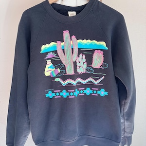 May include: Black sweatshirt with a colorful desert scene featuring cacti, a snake, and geometric patterns. The graphic includes shades of pink, green, blue, and purple. The sweatshirt is hanging on a wooden hanger.