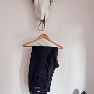 Op de afbeelding: Zwarte denim jeans hangend aan een houten hanger. De jeans hebben een klassiek ontwerp met vijf zakken en een kleine leren patch op de achterzak. De jeans worden gepresenteerd tegen een witte muur, met een koeienschedel en hoorns erboven.