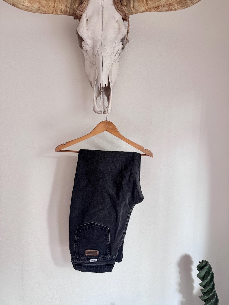 Op de afbeelding: Zwarte denim jeans hangend aan een houten hanger. De jeans hebben een klassiek ontwerp met vijf zakken en een kleine leren patch op de achterzak. De jeans worden gepresenteerd tegen een witte muur, met een koeienschedel en hoorns erboven.