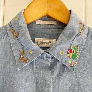 May include: A light blue denim shirt with a white collar and embroidered reindeer and Christmas ornaments. The shirt has a label that reads "Foxcroft" and "100% Cotton Made in Thailand".