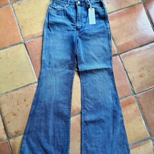 May include: Blue denim flare jeans with a high waist and a button closure. The jeans have a classic five-pocket design and a tag attached to the waistband. The jeans are made of a medium-wash denim fabric.