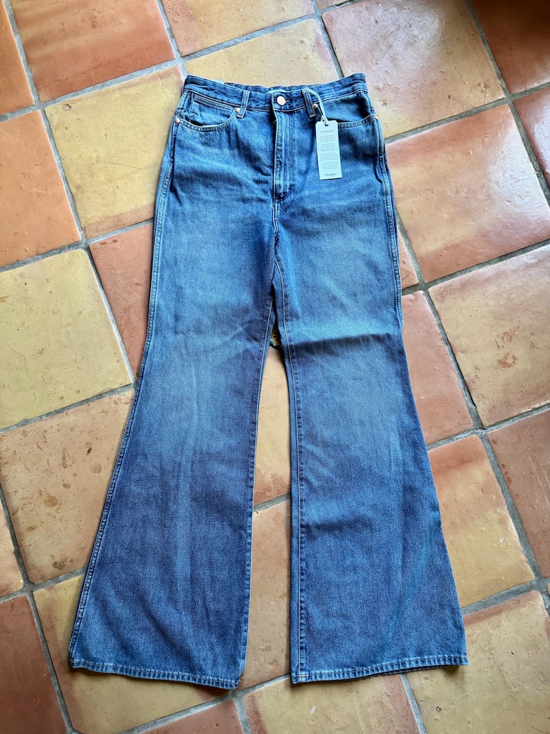 May include: Blue denim flare jeans with a high waist and a button closure. The jeans have a classic five-pocket design and a tag attached to the waistband. The jeans are made of a medium-wash denim fabric.