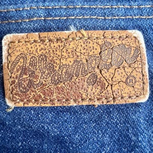 K&ouml;nnte beinhalten: Nahaufnahme eines braunen Lederetiketts mit dem Wort "Mellie" in einer stilisierten Schriftart, auf blauen Denim-Stoff gen&auml;ht. Das Etikett hat ein abgenutztes, rissiges Aussehen. Der Denim hat sichtbare N&auml;hte.