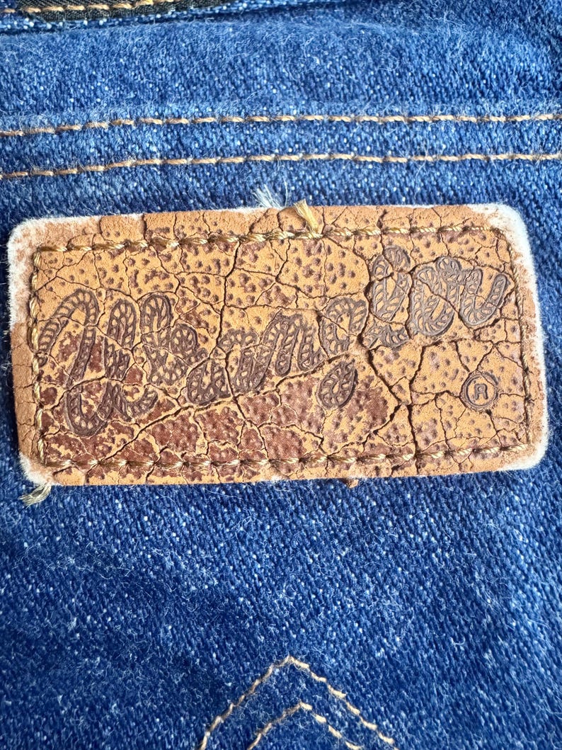 K&ouml;nnte beinhalten: Nahaufnahme eines braunen Lederetiketts mit dem Wort "Mellie" in einer stilisierten Schriftart, auf blauen Denim-Stoff gen&auml;ht. Das Etikett hat ein abgenutztes, rissiges Aussehen. Der Denim hat sichtbare N&auml;hte.