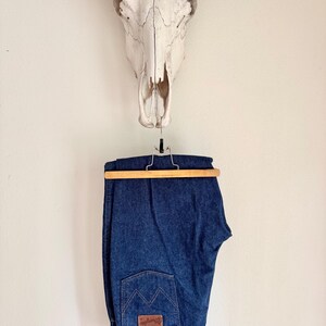 K&ouml;nnte beinhalten: Dunkelblaue Jeans aus Denim, die an einem Holzb&uuml;gel h&auml;ngen. Die Jeans haben ein klassisches F&uuml;nf-Taschen-Design mit einem braunen Lederaufn&auml;her auf der Ges&auml;&szlig;tasche. &Uuml;ber der Jeans ist ein Kuhsch&auml;del an der Wand befestigt.