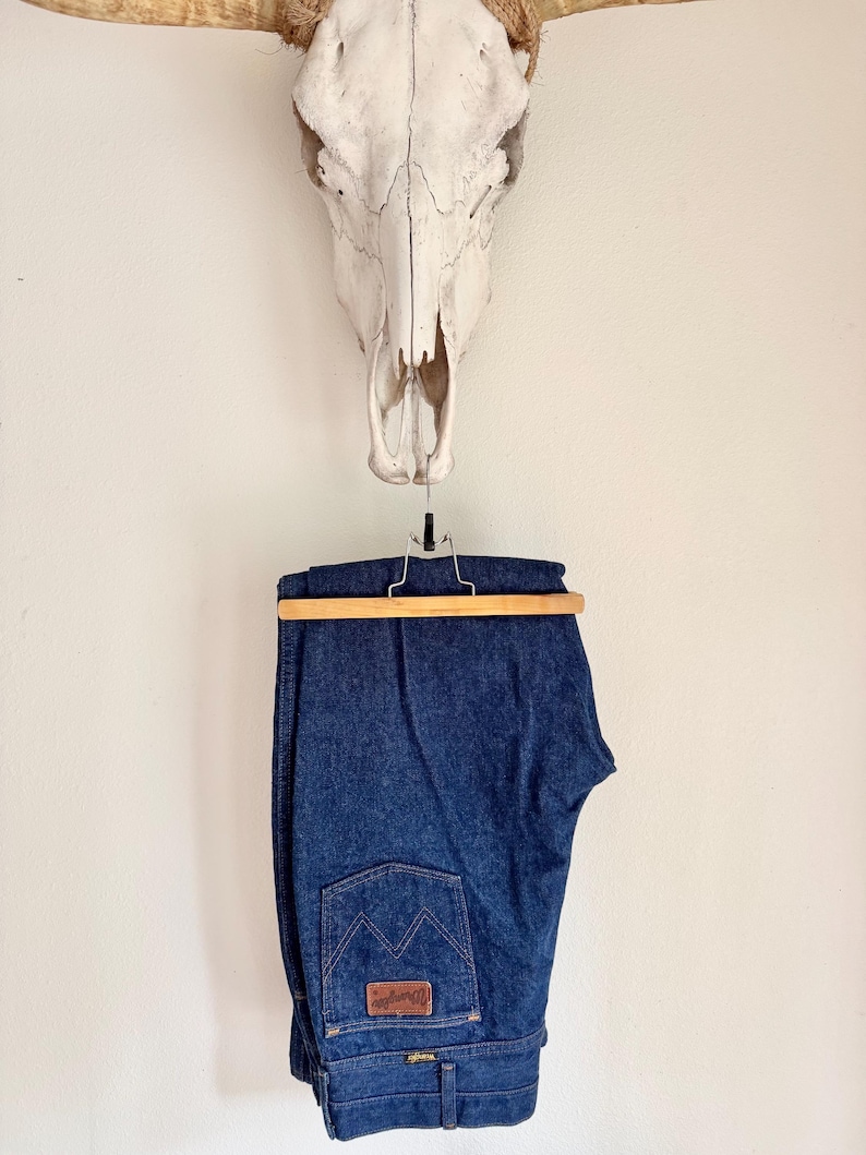 K&ouml;nnte beinhalten: Dunkelblaue Jeans aus Denim, die an einem Holzb&uuml;gel h&auml;ngen. Die Jeans haben ein klassisches F&uuml;nf-Taschen-Design mit einem braunen Lederaufn&auml;her auf der Ges&auml;&szlig;tasche. &Uuml;ber der Jeans ist ein Kuhsch&auml;del an der Wand befestigt.