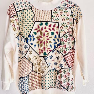 May include: A cream-colored long-sleeve sweatshirt featuring a mock neck and a front panel decorated with a multi-colored geometric patchwork pattern. The design includes various floral and abstract motifs in shades of red, blue, green, and gold.