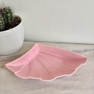 May include: A pink, fan-shaped ceramic dish with a scalloped edge. The dish has a series of vertical ridges, creating a textured surface. A white ceramic pot with a cactus is in the background.