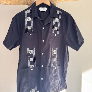 May include: Black short-sleeved shirt with white embroidered details. The shirt features a button-down front, two chest pockets, and decorative embroidery on the front and pockets. The shirt is hanging on a wooden hanger.