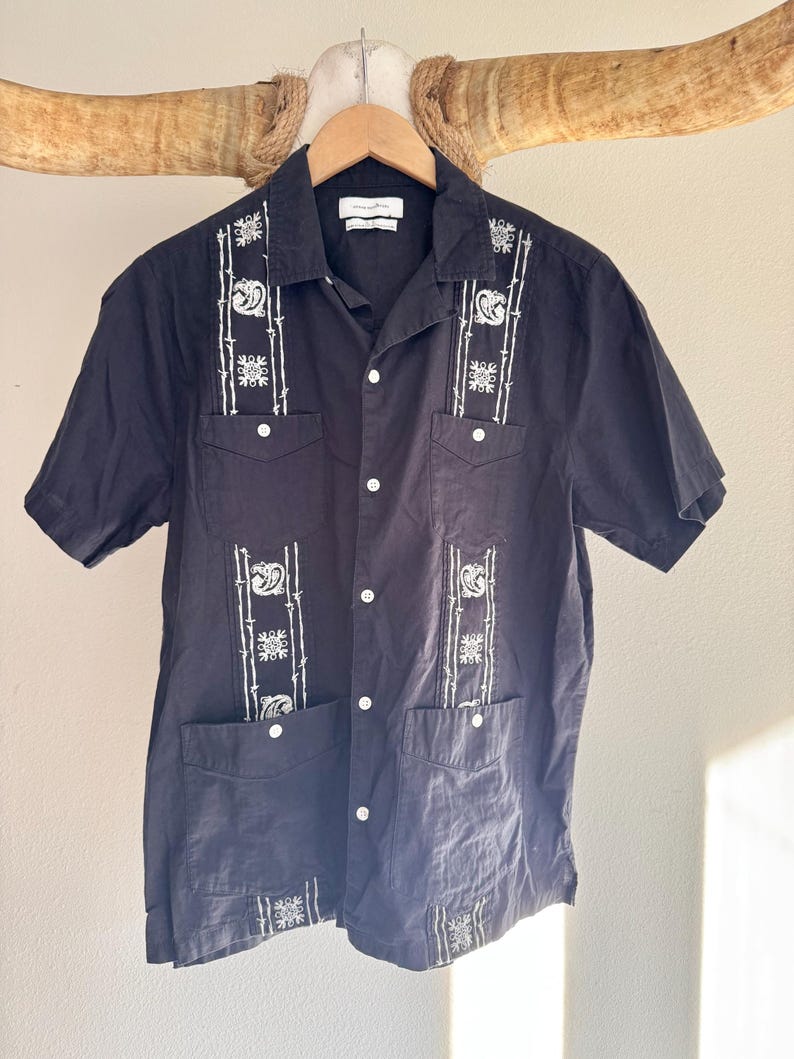 May include: Black short-sleeved shirt with white embroidered details. The shirt features a button-down front, two chest pockets, and decorative embroidery on the front and pockets. The shirt is hanging on a wooden hanger.
