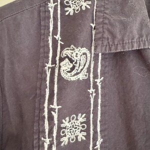 May include: Close-up of a dark navy blue button-down shirt with white embroidered detailing. The embroidery includes floral and paisley designs along the front placket and on the chest pocket.