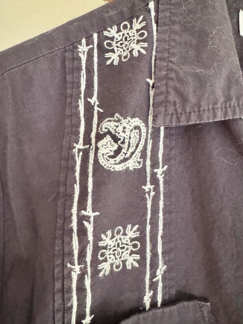 May include: Close-up of a dark navy blue button-down shirt with white embroidered detailing. The embroidery includes floral and paisley designs along the front placket and on the chest pocket.