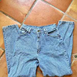 May include: Light blue denim jeans with a classic five-pocket design. The jeans have a high waist and a straight leg cut. The fabric shows signs of wear, giving them a vintage look. The jeans are laid flat on a terracotta tile surface.