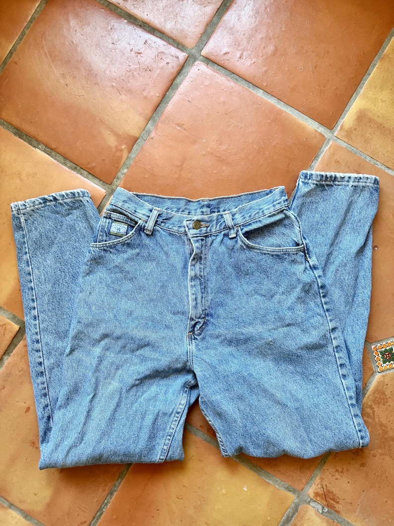 May include: Light blue denim jeans with a classic five-pocket design. The jeans have a high waist and a straight leg cut. The fabric shows signs of wear, giving them a vintage look. The jeans are laid flat on a terracotta tile surface.