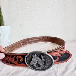 May include: A brown leather belt featuring a black floral-patterned inlay and a large oval silver-tone metal buckle. The buckle displays a raised horse head and horseshoe design. The interior of the belt is stamped with Full Grain Leather U.S.A.