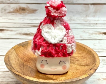 May include: A white ceramic mug with a pink and white crocheted hat with a white heart on the front. The mug has a smiling face with closed eyes and pink cheeks. The mug is sitting on a wooden stand with three legs.