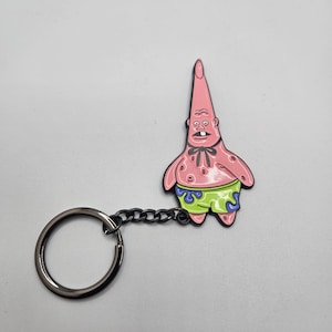 Peut inclure: Un porte-clés représentant un personnage de dessin animé rose, avec une chaîne et un anneau noirs. Le personnage a un corps rose, un short vert et un nœud papillon noir. Sur fond blanc.