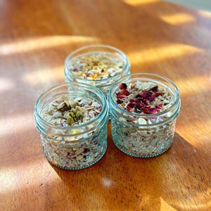 May include: Three glass jars filled with oatmeal and dried flowers. The jars are on a wooden surface. The flowers are pink, yellow, and white.