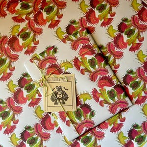 Puede incluir: Papel de regalo blanco con un patrón repetido de plantas de venus atrapamoscas rojas y verdes. El papel está doblado por la mitad y una pequeña etiqueta marrón con el texto "King Arthur Paper Co." está unida al papel.