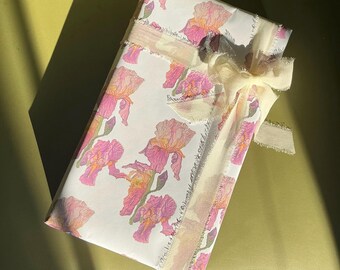 May include: A wrapped gift with white paper featuring a floral iris pattern in pink and orange. The gift is tied with a cream-colored ribbon and bow, creating a delicate and elegant presentation.