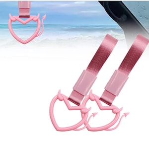 Peut inclure: Charm de téléphone rose en forme de cœur avec des cornes et des queues de diable. Les breloques sont attachées à des sangles en tissu rose. L'arrière-plan montre une image floue de la mer et du ciel.