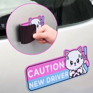 May include: A rectangular car magnet with the text "CAUTION NEW DRIVER" in white on a pink and blue background. A cartoon cat with a worried expression is featured on the right. A hand is shown holding a black magnetic strip.