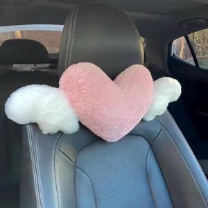 May include: A plush, heart-shaped pillow in a soft pink color, featuring two white, fluffy wing-like attachments on either side. The pillow is positioned against the headrest of a dark gray car seat, showcasing a fuzzy, textured fabric finish.