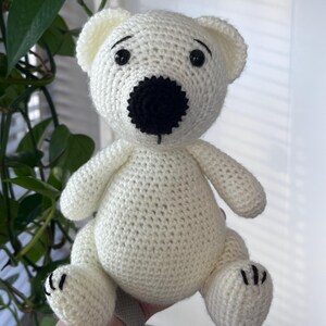 Può includere: Un orsetto all'uncinetto fatto a mano, bianco sporco, con naso e occhi neri. L'orsetto ha piccole orecchie arrotondate ed è seduto in posizione eretta. Le zampe hanno dettagli cuciti neri. È un giocattolo morbido.