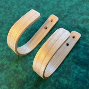 May include: Two light-colored wooden wall hooks with a curved design. Each hook has a rectangular base with two pre-drilled holes for mounting. The hooks are made of a light-colored wood, possibly maple or birch.