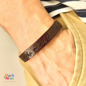 Peut inclure: Un bracelet en cuir marron foncé avec le texte gravé "Live the life you love, Love Always". Le bracelet est porté sur un poignet, avec une chemise rayée et un pantalon kaki en arrière-plan.