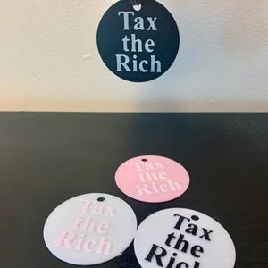 Peut inclure: Trois porte-clés ronds avec le texte "Tax the Rich" en différentes couleurs. Un porte-clés est noir avec du texte blanc, un est blanc avec du texte rose et un est rose avec du texte blanc.