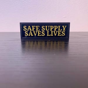 以下が含まれることがあります： 「SAFE SUPPLY SAVES LIVES」と金色で書かれた黒い長方形の看板です。