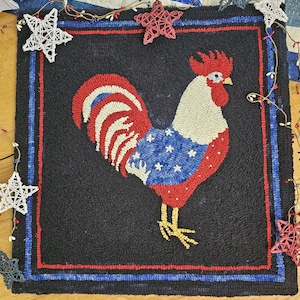 Op de afbeelding: Een zwart tapijt met een haanontwerp in rood, wit en blauw, dat doet denken aan de Amerikaanse vlag. De haan heeft een rode kam en lel, een wit lichaam en een blauwe vleugel met witte sterren. Het tapijt is omlijst met rode en blauwe randen.