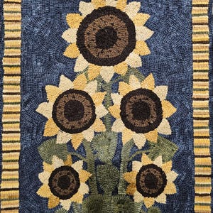 May include: Hand-hooked rug featuring a sunflower design. The rug has a dark blue background with a central arrangement of sunflowers in shades of yellow, brown, and green. A striped border in yellow, brown, and green frames the floral design.