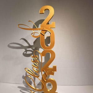 Könnte beinhalten: Eine goldfarbene Skulptur des Jahres "2020" mit dem Wort "Forever" in Schreibschrift. Die Skulptur ist auf einem kleinen rechteckigen Sockel montiert und wirft einen Schatten.