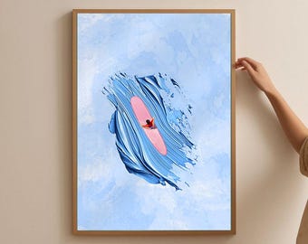 May include: Framed art print featuring a stylized depiction of a surfer on a pink surfboard within a blue wave. The artwork has a textured, brushstroke effect against a light blue background. The frame is a warm, natural wood tone.