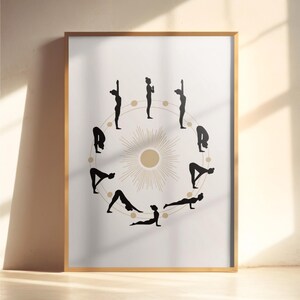Pode incluir: Uma impressão emoldurada com uma ilustração circular de poses de ioga em silhuetas pretas. O design inclui um elemento central de raios de sol em bege e dourado, sobre um fundo branco. A moldura é de cor de madeira clara.