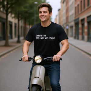 May include: A black t-shirt with the white text "ERROR 404 FEELINGS NOT FOUND". A person is riding a beige scooter on a city street. The person is wearing blue jeans.