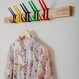 May include: A wooden coat rack with six colorful hooks in black, yellow, green, and red. A floral print dress hangs below on a wooden hanger. The rack is mounted on a white wall.
