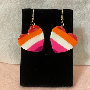 Puede incluir: Un par de pendientes en forma de corazón con un diseño a rayas rojo, naranja, blanco, rosa y morado. Los pendientes están suspendidos de ganchos dorados. Los pendientes se exhiben en un soporte negro sobre un fondo rosa claro.