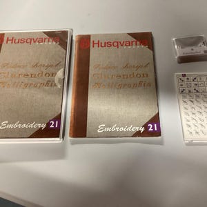 May include: Two Husqvarna Embroidery 21 books with brown and beige covers, and a clear plastic case. The books have gold lettering and a purple corner. Also included are a small clear case and a plastic card with embroidery designs.