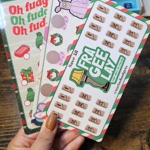 May include: Three colorful sticker sheets with Christmas-themed designs. One sheet has the text "Oh fudg" in red and green, another features a bunny in a pink onesie, and the third says "FRA GEE LAY".
