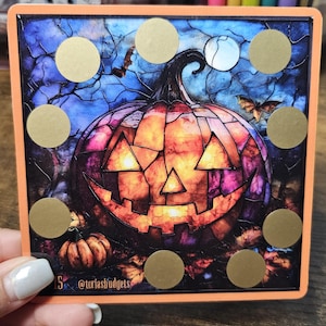 May include: A Halloween-themed sticker sheet featuring a jack-o'-lantern design with a stained-glass effect. The pumpkin is orange, purple, and yellow, with a smiling face. Gold scratch-off circles are arranged around the pumpkin. The text "@toriasbudgets" is visible.