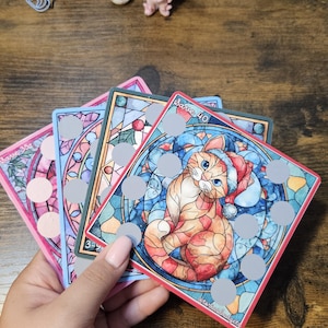 May include: A hand holds four colorful, square cards with cat illustrations in a stained-glass style. Each card features a different cat design, with a festive hat on one. The cards have a pink, blue, and green border.