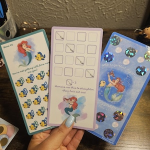 May include: Three colorful cards featuring Ariel from The Little Mermaid. The cards have text and illustrations. One card says "You're not getting cold fins now are you?" Another says "Humans use this to straighten their hair out, see!" The third says "Who says that my dreams?"