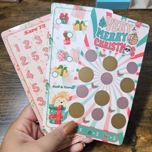 May include: Three Christmas-themed scratch-off cards with festive designs. One card says "Save 75", another says "Roll & Save", and the third says "Very Merry Christmas". The cards feature gift boxes, ornaments, and holly.