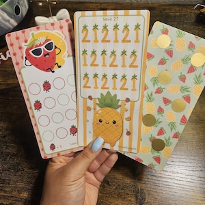 May include: Three colorful budget cards with fruit designs. One card features a strawberry with sunglasses, another has a pineapple, and the third has a pattern of pineapples and strawberries. The pineapple card has the text "Save 27".